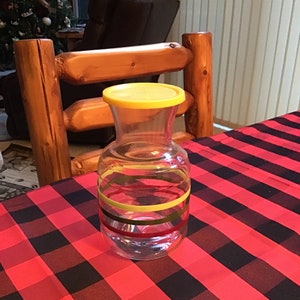 May include: A clear glass pitcher with a yellow lid and striped design. The pitcher is sitting on a red and black checkered tablecloth.