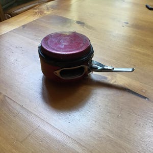 May include: A vintage red and black fishing reel with a silver handle. The reel has a circular design with a textured, maroon-colored top. The handle is attached to the side of the reel.
