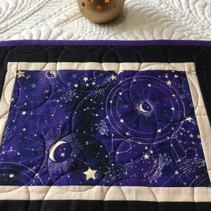 May include: A gold-colored candle holder with star-shaped cutouts sits atop a quilted table runner. The runner features a celestial design with a purple background, white stars, and astrological symbols. The runner is bordered by black and purple fabric.