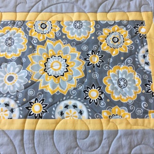 May include: A gray and yellow quilted table runner with a floral pattern. The runner has a yellow border and a gray background with yellow and white flowers.