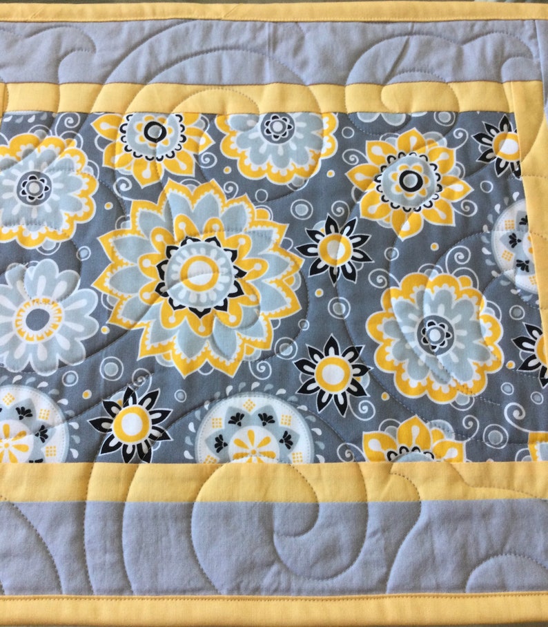 May include: A gray and yellow quilted table runner with a floral pattern. The runner has a yellow border and a gray background with yellow and white flowers.