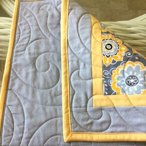 May include: A gray and yellow quilted throw with a swirl pattern. The throw has a yellow border and a floral pattern on the back.