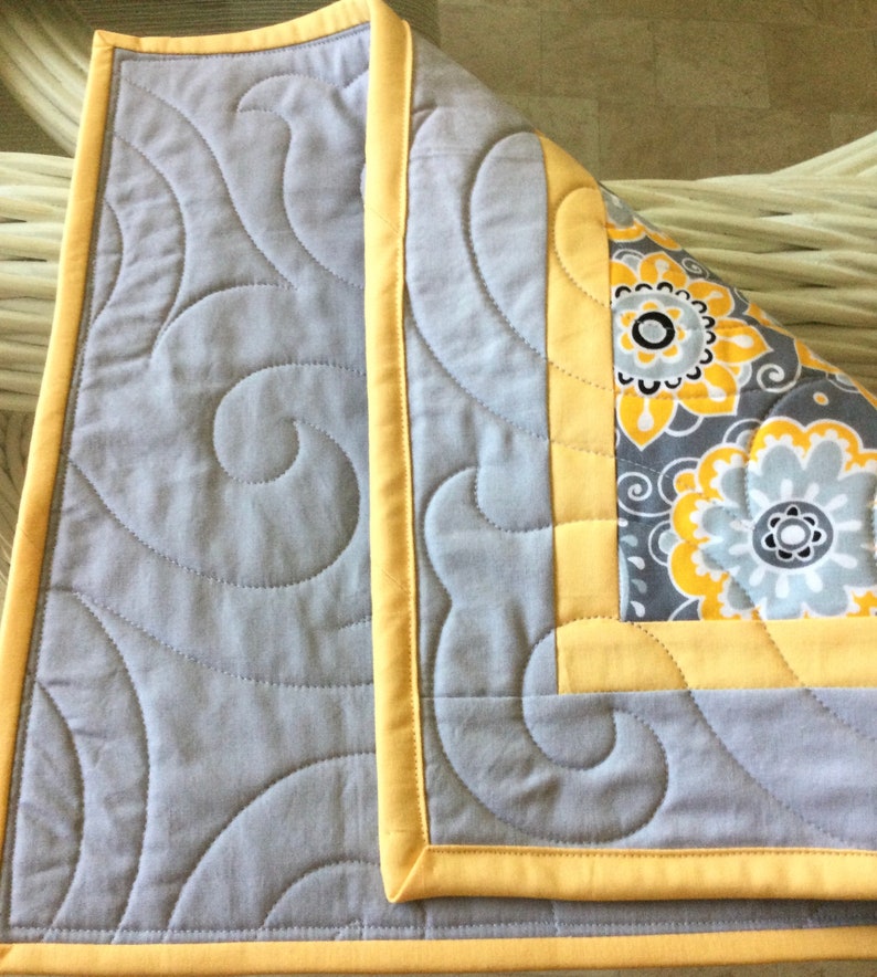 May include: A gray and yellow quilted throw with a swirl pattern. The throw has a yellow border and a floral pattern on the back.