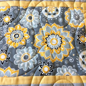 May include: A quilted table runner with a gray background and yellow and white floral pattern. The runner has a yellow border and a quilted design.