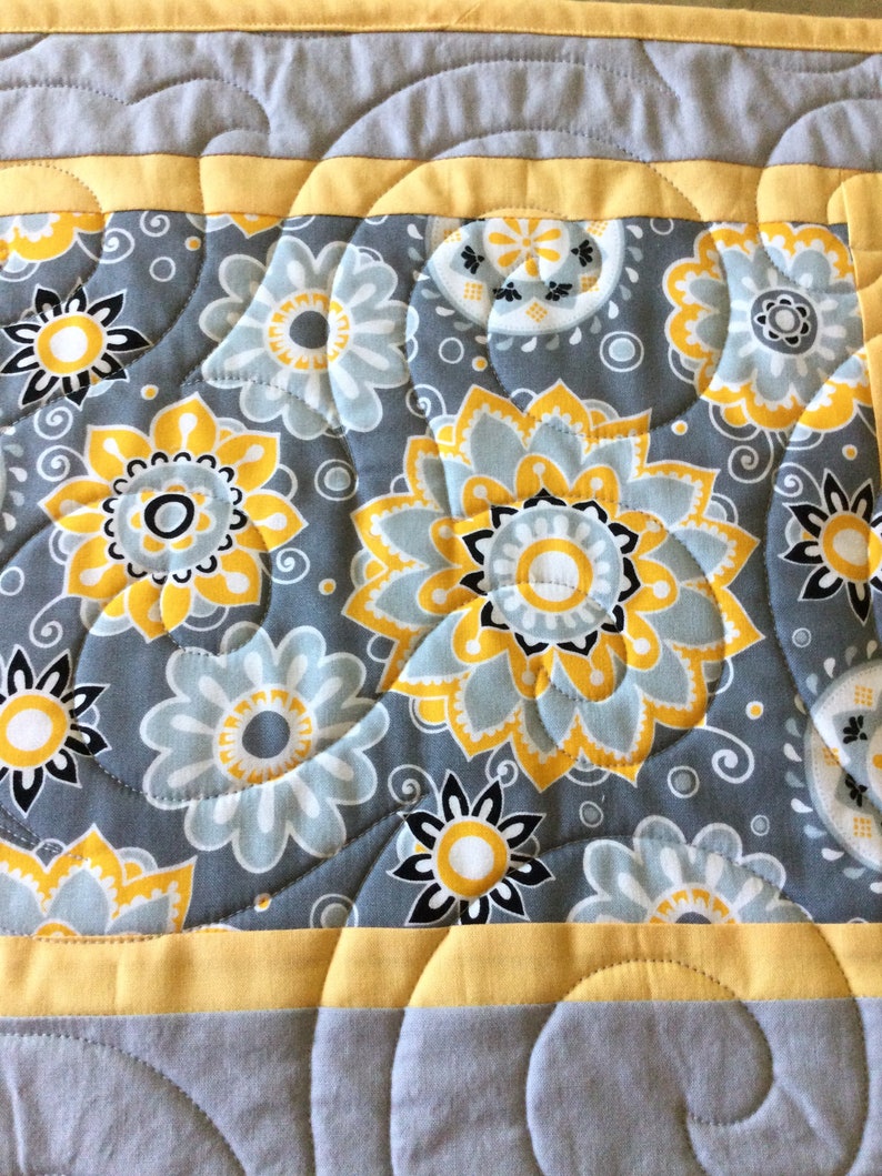 May include: A quilted table runner with a gray background and yellow and white floral pattern. The runner has a yellow border and a quilted design.
