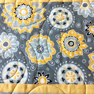 May include: A gray and yellow quilted fabric with a floral pattern. The fabric features a repeating pattern of yellow and white flowers with black centers, surrounded by gray swirls.