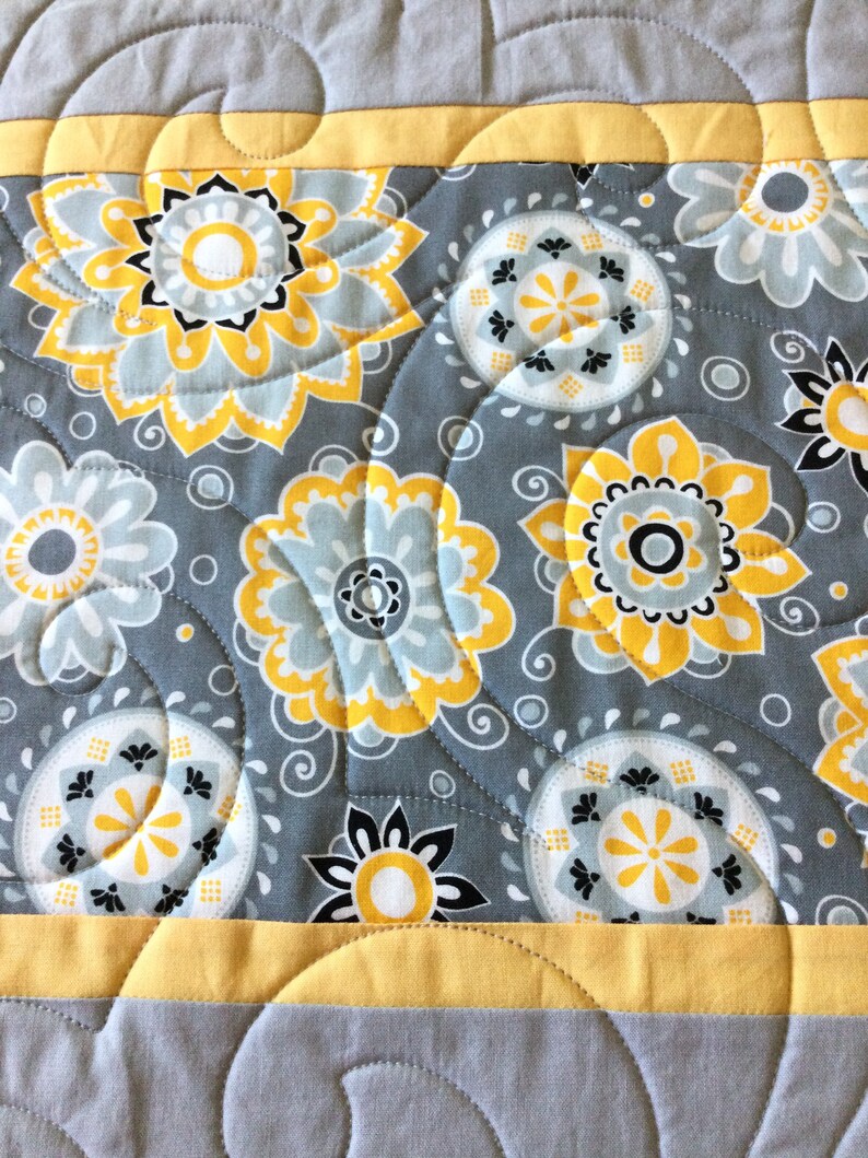 May include: A gray and yellow quilted fabric with a floral pattern. The fabric features a repeating pattern of yellow and white flowers with black centers, surrounded by gray swirls.