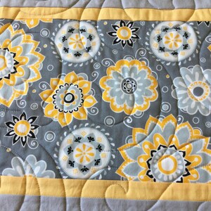May include: A gray and yellow table runner with a floral pattern. The runner has a yellow border and a quilted design.