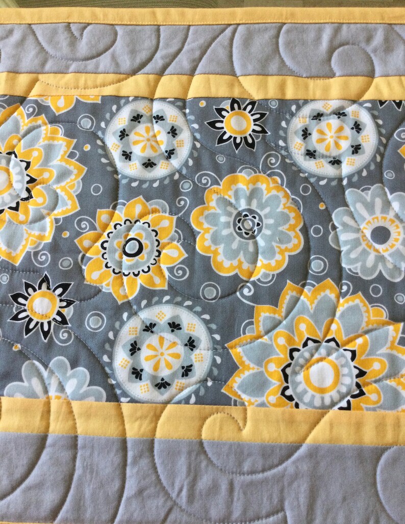 May include: A gray and yellow table runner with a floral pattern. The runner has a yellow border and a quilted design.