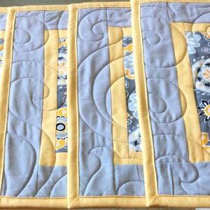 May include: Four gray and yellow quilted placemats with a paisley pattern. The placemats have a yellow border and a gray center with a white and yellow paisley pattern.