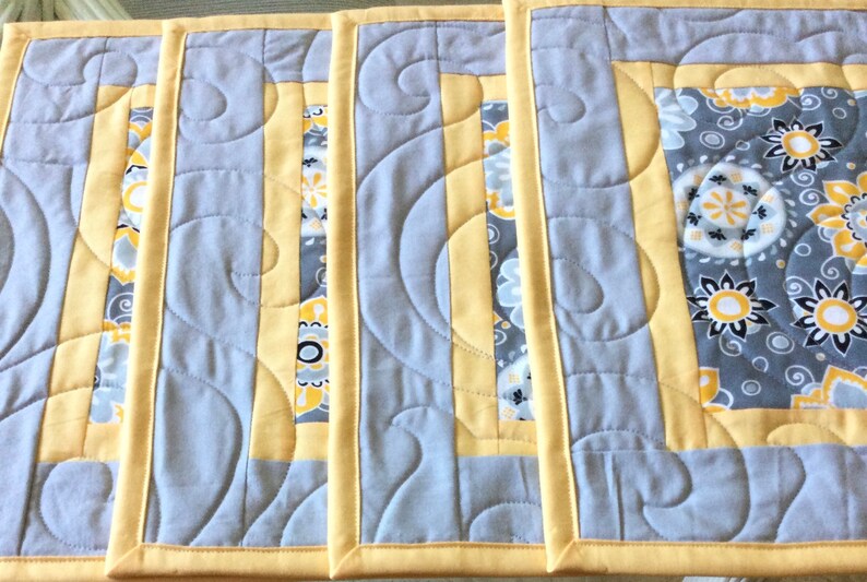 May include: Four gray and yellow quilted placemats with a paisley pattern. The placemats have a yellow border and a gray center with a white and yellow paisley pattern.