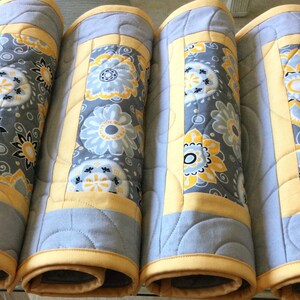 May include: Four gray and yellow quilted placemats with a floral pattern. The placemats are rolled up and ready to be used.