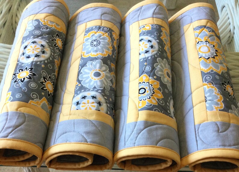 May include: Four gray and yellow quilted placemats with a floral pattern. The placemats are rolled up and ready to be used.