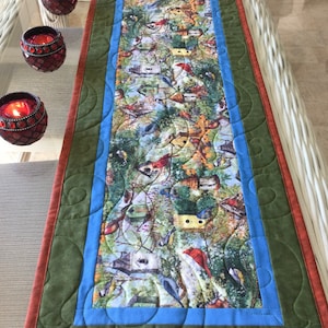 May include: A green, blue, and brown quilted table runner with a bird and birdhouse pattern. The runner has a scalloped edge and a decorative border.