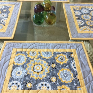 May include: Set of four gray and yellow quilted placemats with a floral pattern. The placemats have a scalloped edge and a yellow border.