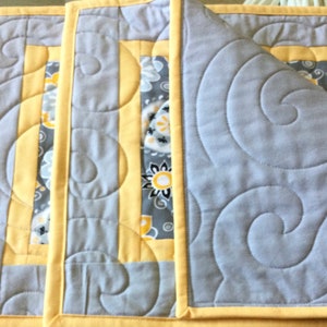 May include: Three gray and yellow quilted placemats with a swirl pattern. The placemats have a yellow border and a gray and white floral pattern in the center.