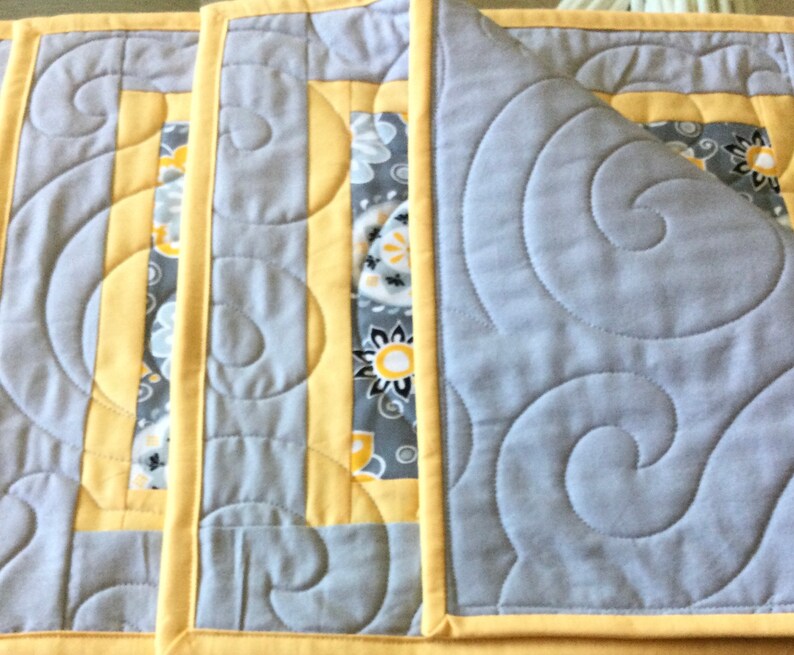 May include: Three gray and yellow quilted placemats with a swirl pattern. The placemats have a yellow border and a gray and white floral pattern in the center.