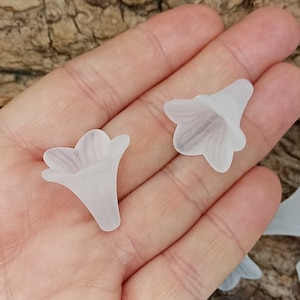 Può includere: Primo piano di una mano che tiene diverse perline di resina bianche a forma di fiore. Le perline hanno un aspetto delicato e traslucido e sono perfette per la creazione di gioielli o altri progetti decorativi.