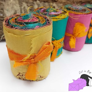 May include: Five rolls of colorful fabric scraps tied with orange ribbon. The fabric scraps are rolled up and tied with a bow. The fabric scraps are in various colors, including yellow, pink, teal, and brown.