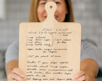 May include: A wooden cutting board with a handle, featuring handwritten recipe instructions for apple crisp. The recipe includes ingredients like oatmeal, brown sugar, and cinnamon. The board is held up by a person.