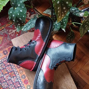 May include: A pair of black and red leather boots with a lace-up design. The boots have a two-tone color scheme, with black on the toe and upper heel, and red on the sides. The boots are displayed on a wooden surface.