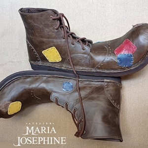May include: A pair of brown leather boots with a unique design. The boots feature colorful patches in yellow, red, and blue. The boots have brown laces and a dark sole. The text "ATELIER MARIA JOSEPHINE" is at the bottom.