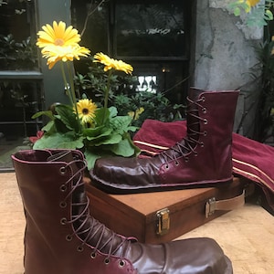 May include: A pair of burgundy leather boots with brown leather toes and laces. The boots are sitting on a wooden surface with a wooden box behind them.