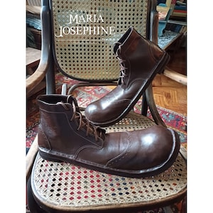May include: Brown leather boots with lace-up closures and decorative perforations. The boots are displayed on a wicker chair. The image includes the text "MARIA JOSEPHINE".