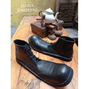 May include: A pair of black leather ankle boots with a rounded toe and lace-up closure. The boots are displayed on a wooden surface. A vintage teapot and a wooden figure are in the background.