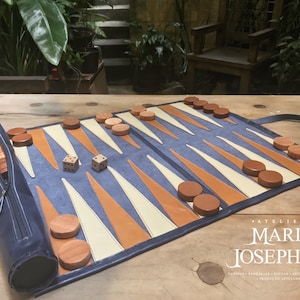 May include: A blue leather backgammon set with wooden playing pieces and dice. The set is rolled up and has a zippered pouch. The set is on a wooden table with the text "Atelier Maria Josephine" visible.