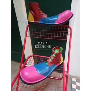 May include: A pair of colorful clown shoes with a red, yellow, blue, and pink color scheme. The shoes are made of leather and have a pointed toe. The shoes are sitting on a red metal chair. The text "Atelier Maria Josephine" is visible on the chair.