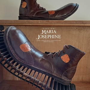 May include: Brown leather boots with a unique design, featuring a rounded toe and a patch detail. The boots are displayed on a wooden surface. The image includes the text "ATELIER MARIA JOSEPHINE".