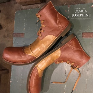 May include: A pair of two-tone leather boots. The boots are brown and tan with a lace-up design. The boots have a rounded toe and a low heel. The boots are sitting on a weathered green surface.