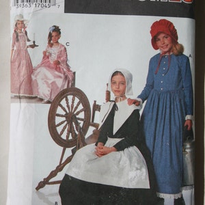 May include: Simplicity Costumes pattern featuring historical children's outfits. The package shows illustrations of girls in colonial-era dresses, bonnets, and a spinning wheel. The text "Simplicity Costumes" is displayed in red.
