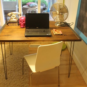 May include: A wooden desk with hairpin legs, a laptop computer, a pink plastic skull, a vintage rotary phone, and a silver fan. The desk is set against a white wall with a black chalkboard with a blue frame.