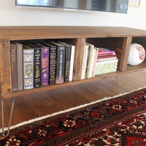May include: A rustic wooden media console with three open shelves, featuring a collection of books and a white volleyball. The console is supported by four hairpin legs.