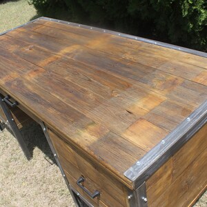 May include: A rustic wooden desk with metal accents. The desk has a large top surface and two drawers. The drawers have metal handles. The desk is made of reclaimed wood and has a distressed finish.