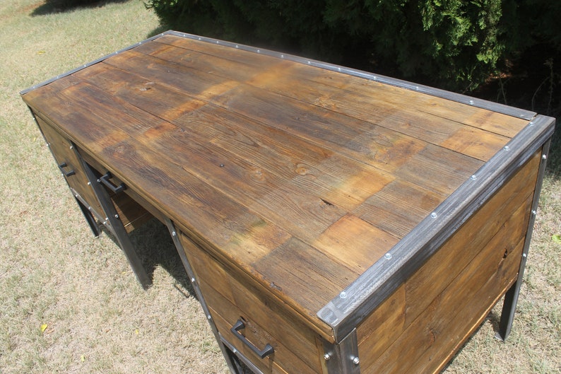 May include: A rustic wooden desk with metal accents. The desk has a large top surface and two drawers. The drawers have metal handles. The desk is made of reclaimed wood and has a distressed finish.