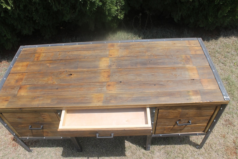 May include: A rustic wooden desk with metal accents and three drawers. The desk has a distressed wood finish and metal hardware. The top drawer is open.