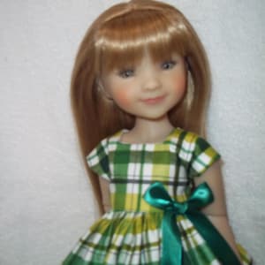 May include: A doll with long, light brown hair and a green and white plaid dress. The dress features a green satin bow at the waist. The doll has rosy cheeks and is smiling.