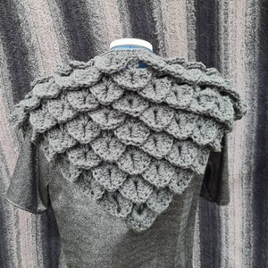 May include: Grey crocheted shawl with a scale pattern, worn over a grey shirt.