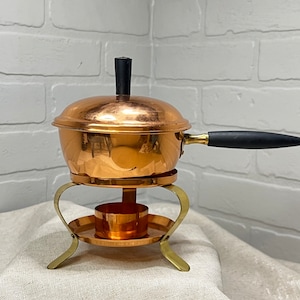 May include: A copper fondue pot with a black handle and lid knob. The pot sits on a brass stand with a small burner cup. The pot is designed for melting cheese or chocolate.