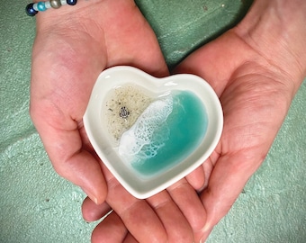 May include: A heart-shaped white ceramic dish with a beach scene. The dish features a sandy area with a small turtle figurine and a turquoise resin ocean with white foamy waves. The dish is held in open hands.