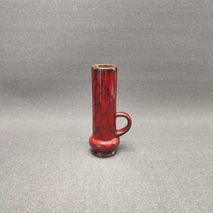 Puede incluir: Un pequeño jarrón de cerámica rojo con asa. El jarrón tiene un esmalte rojo y negro moteado.
