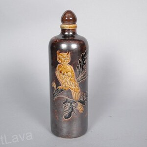 May include: A brown ceramic bottle with a cork stopper and a hand-painted design of an owl perched on a branch. The owl is depicted in shades of brown and yellow.