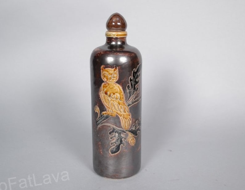 May include: A brown ceramic bottle with a cork stopper and a hand-painted design of an owl perched on a branch. The owl is depicted in shades of brown and yellow.