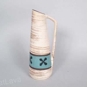 Könnte beinhalten: Eine weiße Keramikvase mit braunem und blauem Streifenmuster und schwarzen Kreuzakzenten. Die Vase hat einen Henkel und eine schmale Öffnung.