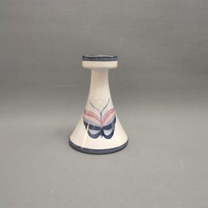 May include: A white ceramic candle holder with a blue and pink butterfly design. The holder has a flared base and a narrow top.