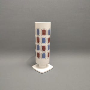 Puede incluir: Jarrón de cerámica blanco con base cuadrada. El jarrón tiene un cuerpo cilíndrico con un patrón repetitivo de cuadrados azules y rojos con diseños geométricos.
