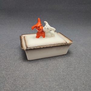 May include: A white ceramic trinket box with a brown rim and a lid featuring two small ceramic dogs. One dog is orange and the other is white.